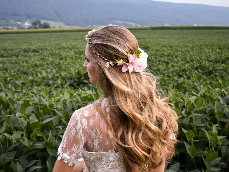 Puede incluir: Una mujer con cabello largo y ondulado rubio, adornado con una corona floral y una flor rosa. Viste un vestido de encaje blanco, de pie en un campo de plantas verdes con una cordillera al fondo. La imagen captura una est&eacute;tica natural y rom&aacute;ntica.