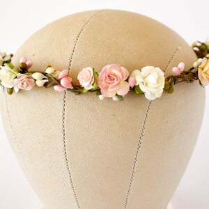 Op de afbeelding: Een bloemenkrans met roze, witte en perzikrozen, knoppen en groen. De bloemen zijn gerangschikt op een flexibele, bruine draadbasis. De kroon wordt gepresenteerd op een mannequinhoofd.