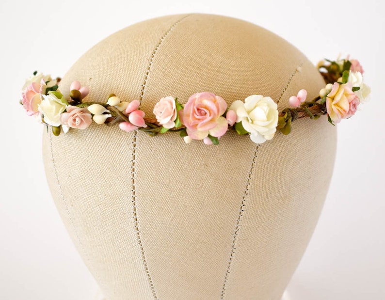 Op de afbeelding: Een bloemenkrans met roze, witte en perzikrozen, knoppen en groen. De bloemen zijn gerangschikt op een flexibele, bruine draadbasis. De kroon wordt gepresenteerd op een mannequinhoofd.