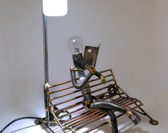 May include: A metal sculpture of a person sitting on a bench reading a book. The sculpture is made of various metal parts, including nuts, bolts, and pipes. The person has a light bulb for a head and is holding a metal book. The sculpture is illuminated by a lamp with a white shade.