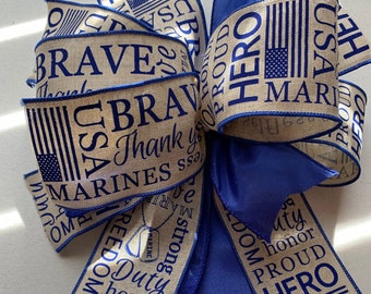 May include: A patriotic bow featuring tan ribbon printed with blue text and American flags. The text includes "BRAVE", "HERO", "USA", "MARINES", and "Thank you". The bow has a vibrant blue satin ribbon accent.