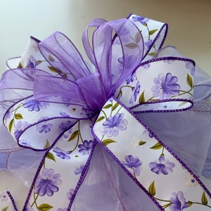 May include: A purple and white floral ribbon bow with a sheer purple ribbon in the center. The bow is tied with a purple satin ribbon.