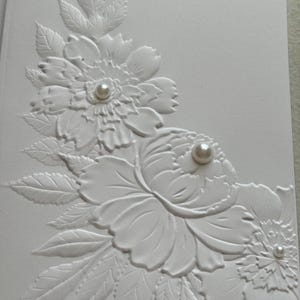 May include: A white card with embossed floral designs. The card features detailed relief patterns of flowers and leaves, with pearl embellishments at the center of each flower. The overall design is elegant and suitable for various occasions.