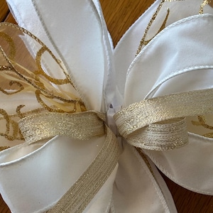 May include: A large, decorative bow featuring wide, white satin ribbons and gold, sheer ribbons with a swirling pattern. The bow is tied with a gold ribbon, creating a festive and elegant look.