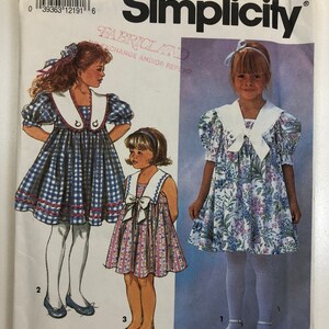 May include: Simplicity sewing pattern number 7697 for girls' dresses. The pattern includes two dress styles: a gingham dress with a white collar and a floral dress with a white collar. The pattern is for sizes AA (2-4).