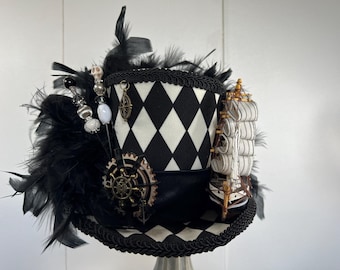 May include: A black and white harlequin top hat with a diamond pattern, adorned with black feathers, decorative beads, and a miniature ship. The hat features a gear and compass embellishment, creating a steampunk aesthetic.
