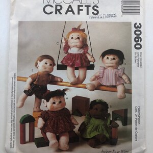 Op de afbeelding: McCall's Crafts naaipatroon envelop met stoffen poppen in verschillende outfits, met houten blokken en een schommel. De envelop bevat de tekst "FANNY'S FABRICS" en "by/par Faye Wine".