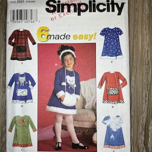 Może przedstawiać: Koperta z wykrojem do szycia Simplicity z ilustracjami sukienek dla dzieci. Koperta jest biała z logo Simplicity i napisem "6 made easy!". Sukienki są w różnych kolorach i wzorach, w tym w kratę, gwiazdy i wzór misia.