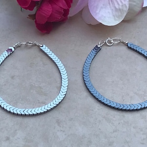 Peut inclure: Deux bracelets en hématite avec un motif en chevrons. Les bracelets sont argentés et gris foncé, avec une fermeture à fermoir homard. Ils sont présentés sur une surface claire, avec des fleurs roses et blanches en arrière-plan.