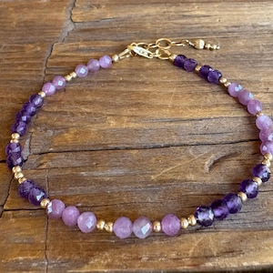 May include: A delicate bracelet featuring amethyst and gold beads. The bracelet alternates between faceted purple and lavender beads, connected by small gold-colored beads and a gold clasp. The jewelry is displayed on a rustic wooden surface.