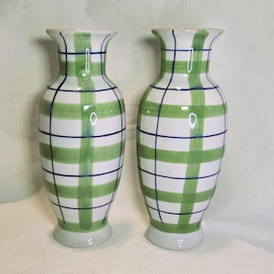 May include: Two matching ceramic vases. Each vase is white with a green plaid pattern and dark blue grid lines. The vases have a flared top and a round base. The vases are decorative.