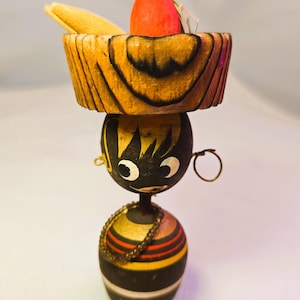 May include: A whimsical wooden figurine of a person with a sombrero. The figurine has a black face with white eyes and a necklace. The sombrero is brown with a red object and a yellow object inside.