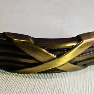 May include: A bronze-toned cabinet pull with a unique design. The handle features a series of horizontal lines crossed by a decorative, interwoven element. The hardware has a vintage aesthetic.