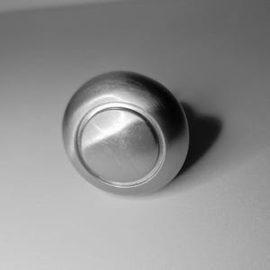 May include: A close-up shot of a brushed metal knob. The knob is round with a flat, circular face and a slightly domed body. The surface has visible grain and a metallic sheen, reflecting light. The object is set against a neutral background.