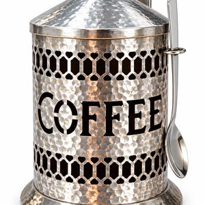 May include: A cylindrical silver-toned metal coffee canister with a hammered texture, a removable lid with a knob, and a decorative cutout pattern. The word COFFEE is printed in black letters on the front, and a matching metal spoon is included.