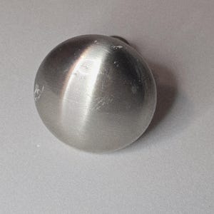 May include: A brushed metal sphere with a small, dark attachment at the top. The surface of the sphere has visible scratches and imperfections. The object is set against a light gray background.