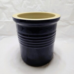 May include: A deep navy blue ceramic crock with a wide, flared rim and a smooth, glossy finish. The exterior features a series of horizontal, recessed ridges encircling the upper portion of the vessel, while the interior is a light cream color.