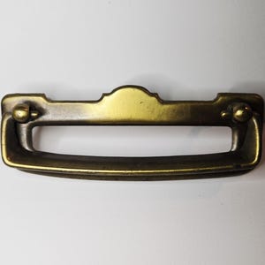 May include: A vintage-style brass drawer pull with a curved handle and decorative backplate. The pull features a warm, golden hue with subtle variations in tone, suggesting age and use. The handle is approximately 5 inches long.
