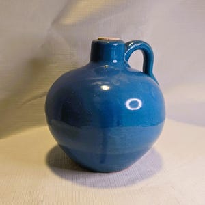 May include: A blue ceramic jug with a round body, a handle, and a small opening at the top. The jug has a cork stopper and a glossy finish. The jug is a deep shade of blue.