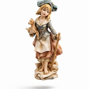 May include: A detailed figurine of a young girl dressed in a blue hat and jacket, holding a lamb. She stands on a cloud-like base, with a wooden staff in her right hand. The figurine is painted in soft, muted colors.