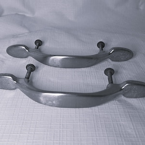May include: Two silver-toned metal drawer pulls. Each pull has a curved handle with oval end pieces and two screw posts for mounting. The pulls are likely for a cabinet or drawer.