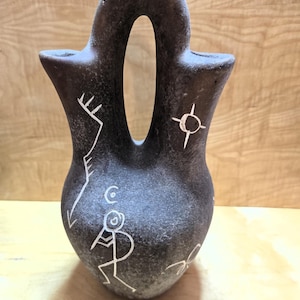 May include: A dark gray, textured double-spout vessel with a central arched handle. The surface features white etched line drawings, including a central figure playing a flute, a sun-like symbol, and geometric patterns across the rounded body.