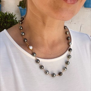 May include: A gold chain necklace with black and white pearls. The necklace is on a person wearing a white shirt.
