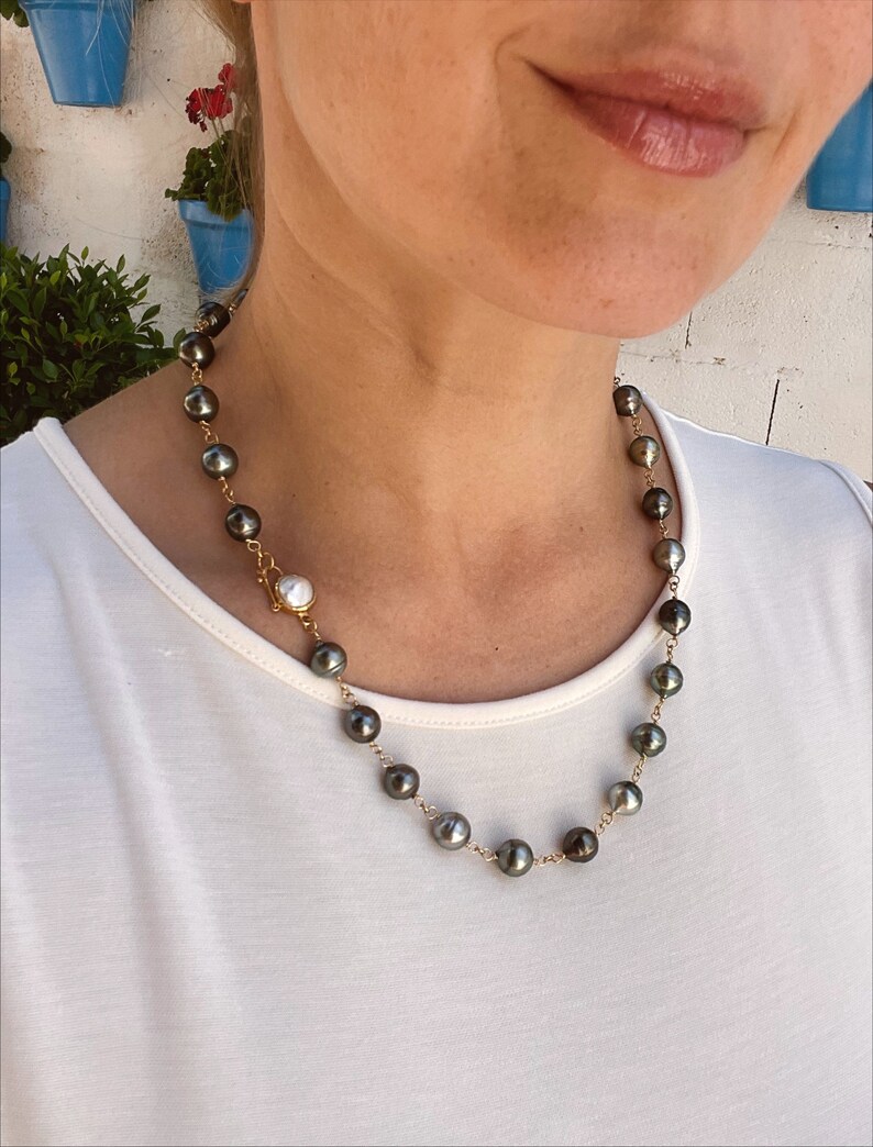 May include: A gold chain necklace with black and white pearls. The necklace is on a person wearing a white shirt.
