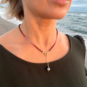May include: A delicate necklace with a single black pearl pendant. The necklace is made of small, pink and red beads and has a gold clasp.