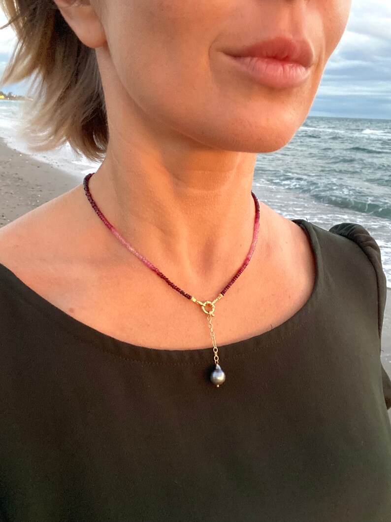 May include: A delicate necklace with a single black pearl pendant. The necklace is made of small, pink and red beads and has a gold clasp.
