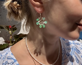 Puede incluir: Un par de pendientes de oro con un racimo de piedras preciosas verdes y pequeñas perlas blancas. Los pendientes tienen forma de flor.