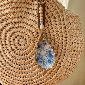 May include: A round woven straw handbag with a brown leather handle. A decorative charm hangs from the handle, featuring wooden beads, a pearl, and a blue floral design on an oyster shell with a gold trim.