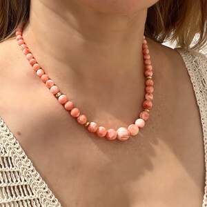 May include: A delicate necklace made of pink coral beads with gold accents. The necklace is worn by a person with a light complexion.