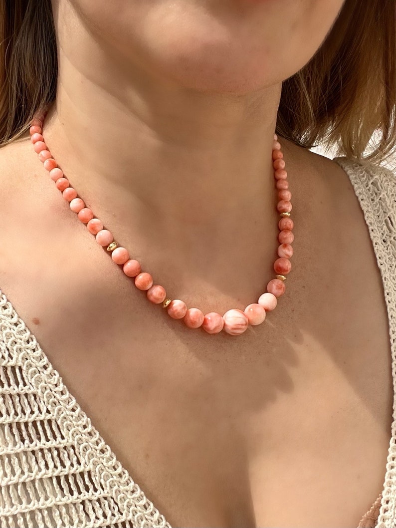 May include: A delicate necklace made of pink coral beads with gold accents. The necklace is worn by a person with a light complexion.