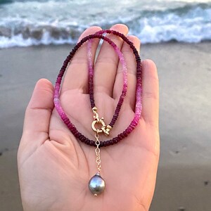 May include: A pink to red ombre gemstone necklace with a gold clasp and a single grey pearl pendant.
