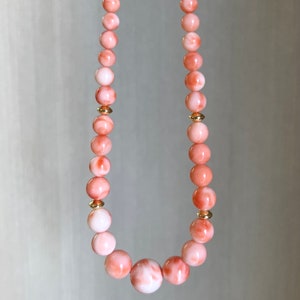 May include: A pink and white coral bead necklace with gold accents. The necklace is made of round beads and has a larger, round bead in the center.