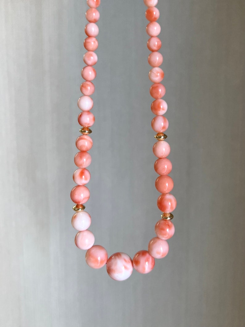 May include: A pink and white coral bead necklace with gold accents. The necklace is made of round beads and has a larger, round bead in the center.