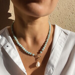 Può includere: Una collana di perle blu chiaro con una chiusura dorata e un grande ciondolo di perla bianca.