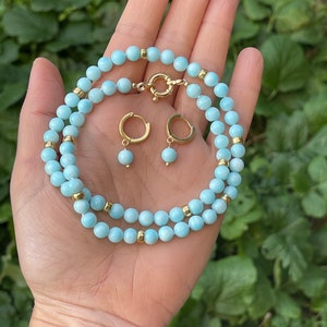 Op de afbeelding: Een lichtblauwe edelsteen ketting en bijpassende oorbellen. De ketting is gemaakt van kleine, ronde kralen met gouden accenten. De oorbellen zijn eenvoudige hoepels met een enkele kraal die aan elke hangt.