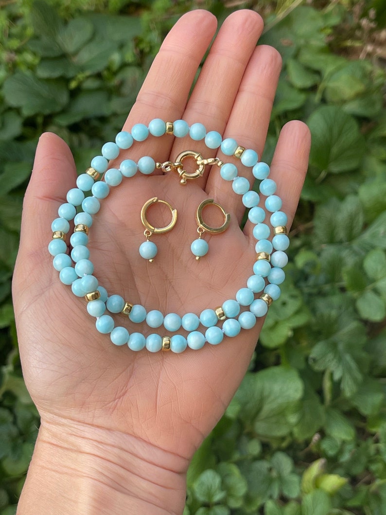 Op de afbeelding: Een lichtblauwe edelsteen ketting en bijpassende oorbellen. De ketting is gemaakt van kleine, ronde kralen met gouden accenten. De oorbellen zijn eenvoudige hoepels met een enkele kraal die aan elke hangt.