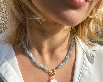 Puede incluir: Un collar delicado con una cadena de cuentas de piedras preciosas azul claro y un gran colgante de perla barroca blanca. El collar tiene un cierre de oro y lo lleva una persona con el pelo rubio.