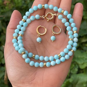 Op de afbeelding: Een lichtblauwe edelsteenketting met gouden accenten en bijpassende oorbellen. De ketting is gemaakt van ronde kralen en heeft een sluiting. De oorbellen zijn hoepels met een hangende edelsteen.