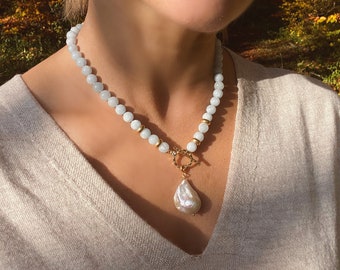 Puede incluir: Un collar de cuentas azul claro con un gran colgante de perla blanca. El collar está hecho de pequeñas cuentas redondas y tiene un cierre dorado.