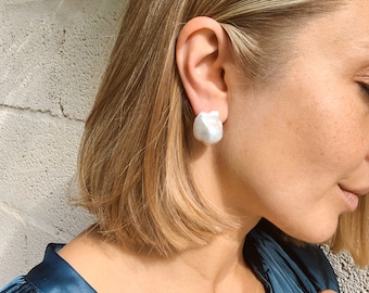 May include: A pair of large, white, baroque pearl earrings. The pearls are a soft, off-white color and have an irregular shape. The earrings are worn by a person with blonde hair.