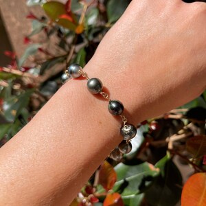 May include: A gold chain bracelet with dark gray pearls. The bracelet is on a person's wrist.
