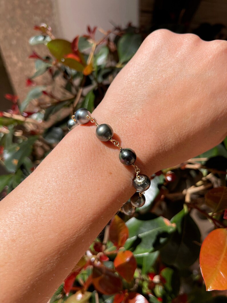 May include: A gold chain bracelet with dark gray pearls. The bracelet is on a person's wrist.