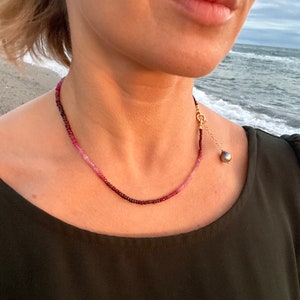 May include: A delicate necklace made of small pink and red gemstones. The necklace has a gold clasp and a single black pearl pendant.
