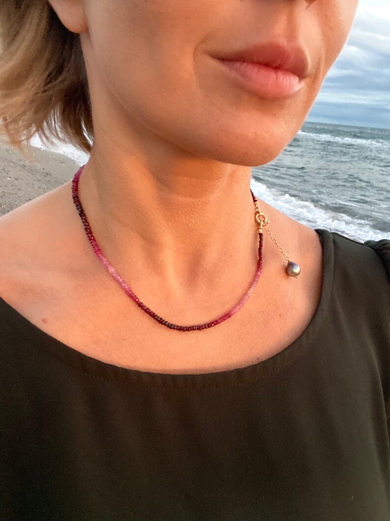 May include: A delicate necklace made of small pink and red gemstones. The necklace has a gold clasp and a single black pearl pendant.