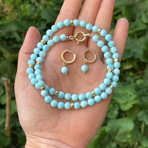 Op de afbeelding: Een lichtblauwe edelsteen ketting en bijpassende oorbellen. De ketting is gemaakt van kleine, ronde kralen en heeft een gouden sluiting. De oorbellen zijn ook gemaakt van kleine, ronde kralen en hebben gouden hoepels.