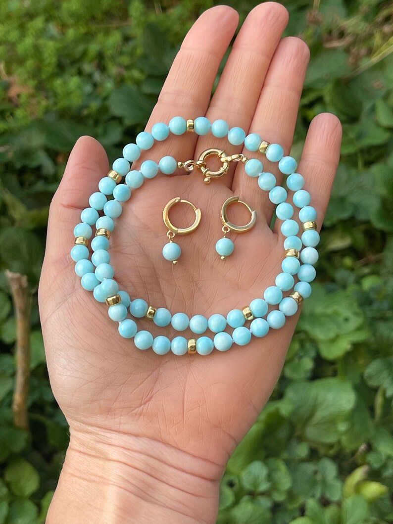 Op de afbeelding: Een lichtblauwe edelsteen ketting en bijpassende oorbellen. De ketting is gemaakt van kleine, ronde kralen en heeft een gouden sluiting. De oorbellen zijn ook gemaakt van kleine, ronde kralen en hebben gouden hoepels.